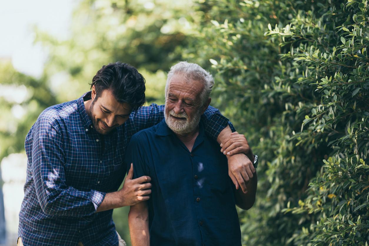 An adult son walks with his older father through a park with an arm around his shoulders.